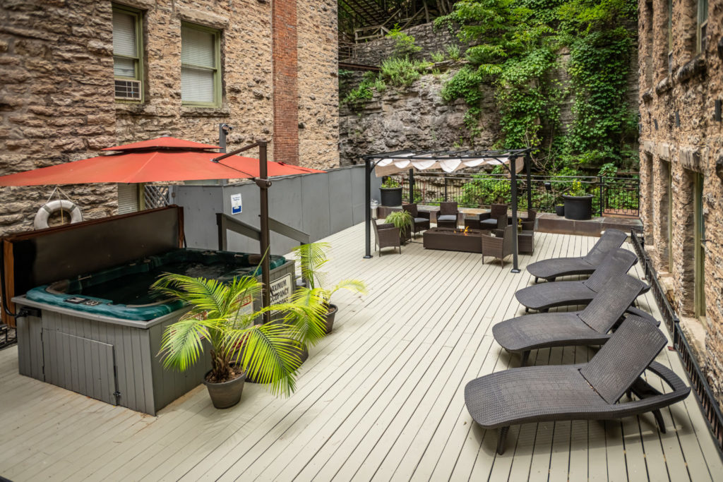 Sun deck tucked between two historical brick buildings. Several plants, a hot tub, and lounge chairs cover the deck.