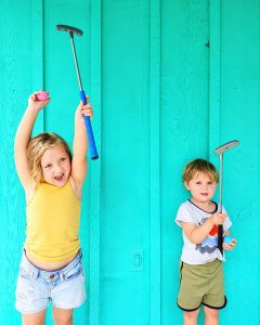 A young girl and boy holding golf clubs against a bright turquoise wooden wall in Eureka Springs, Ar.