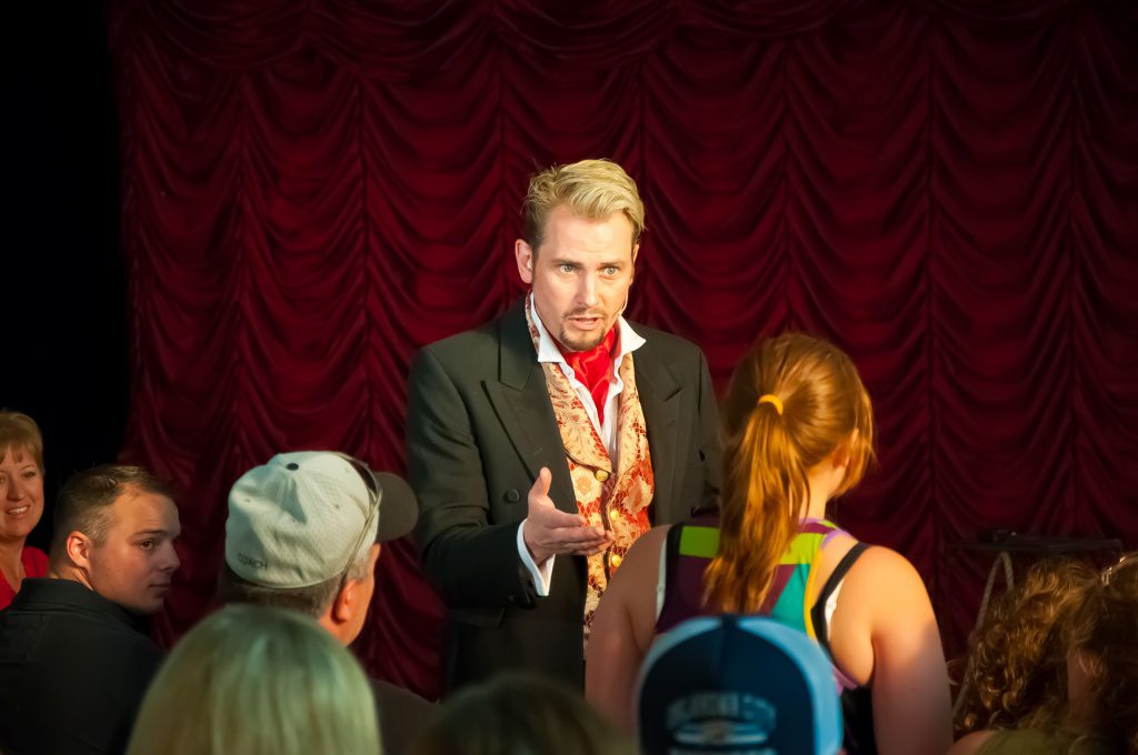 A performer speaking to an audience on stage with red curtains in the background.